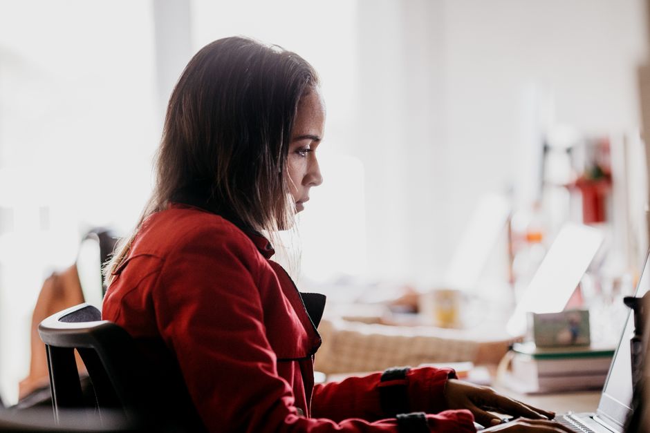 Focused young woman in red jacket typing on laptop in bright workspace.